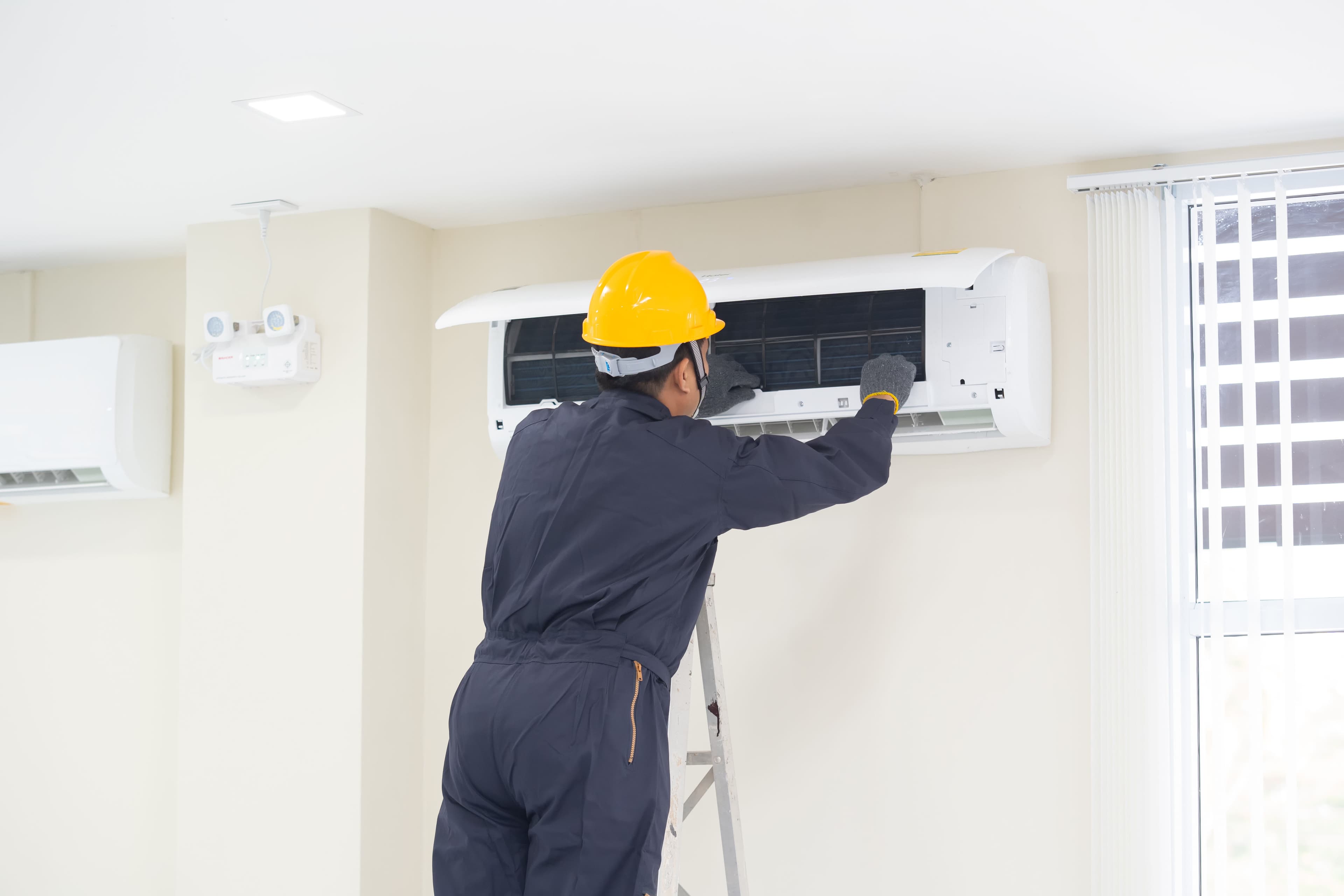 Technician in hard hat servicing wall-mounted air conditioner unit.