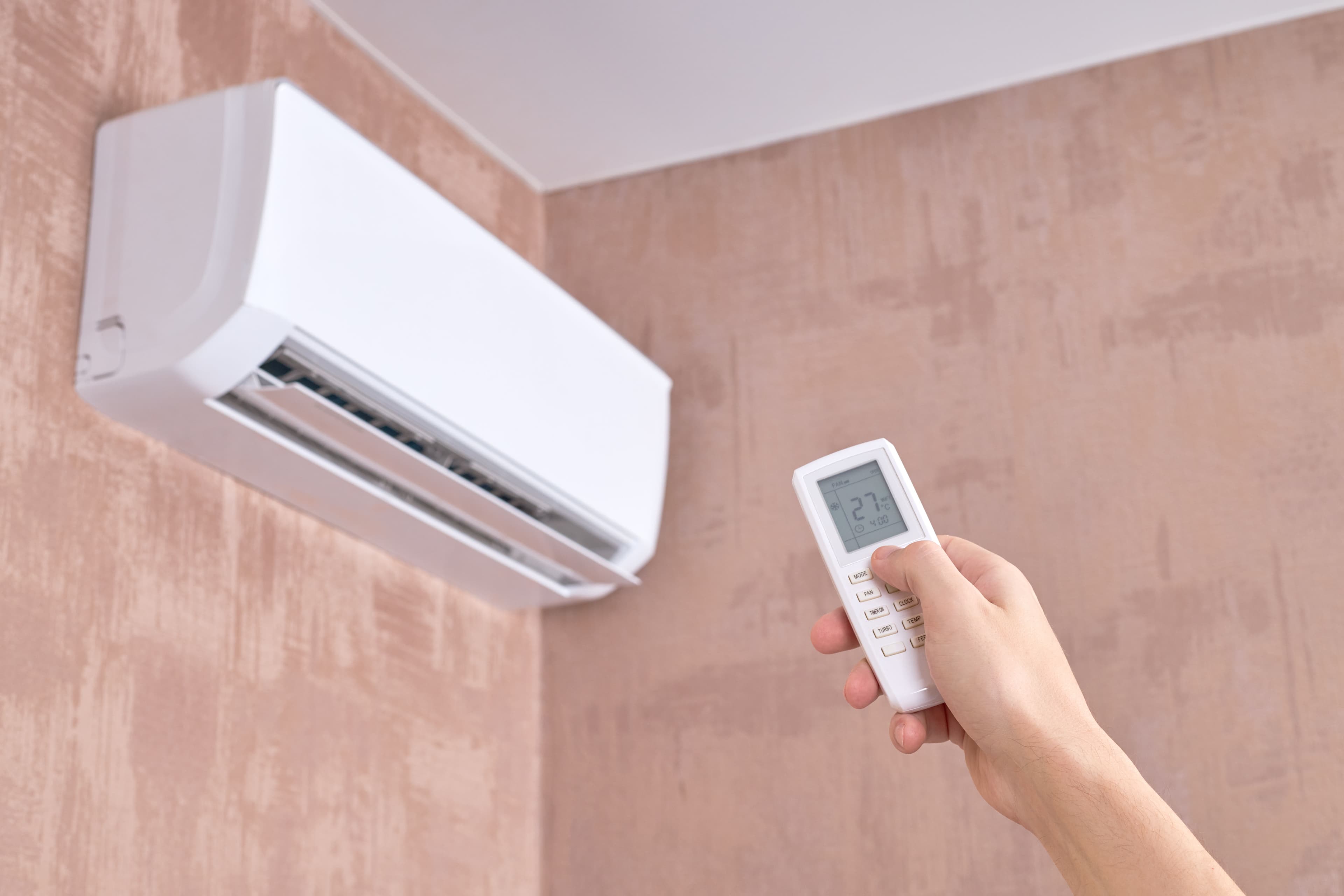 Person adjusting air conditioner temperature with remote control on beige wall.