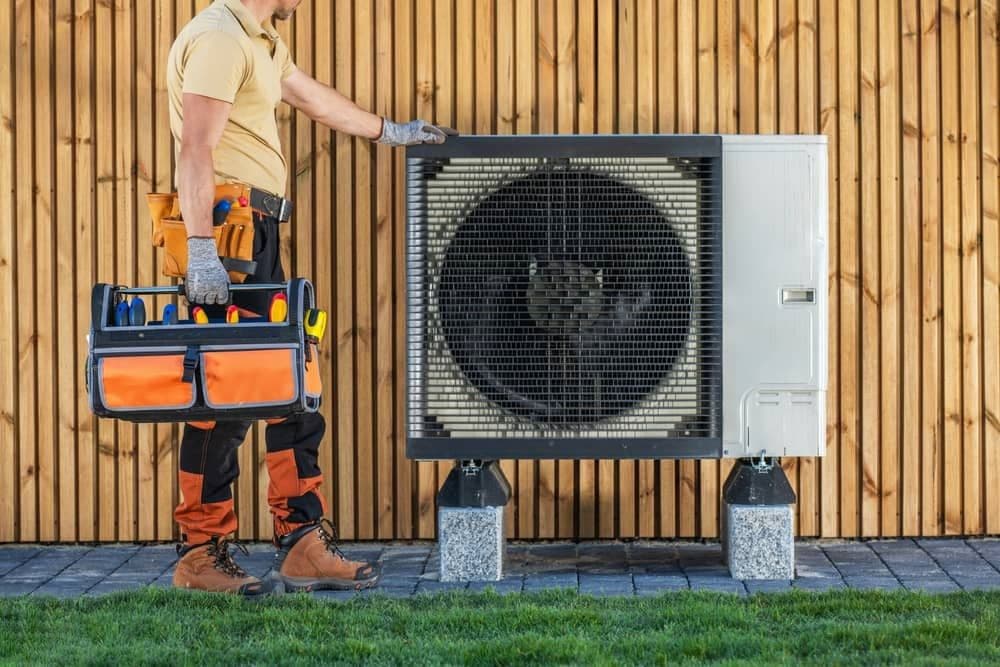 HVAC technician adjusting outdoor air conditioning unit beside tool bag on the ground.