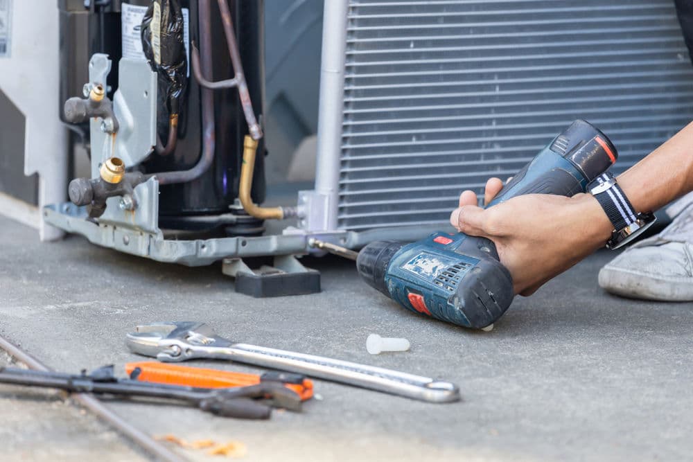 Person using a power drill to repair an appliance with tools nearby on a concrete surface.