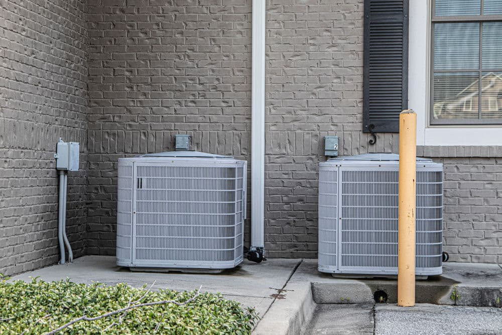 Two outdoor air conditioning units beside a beige brick wall with a window and utility connection.