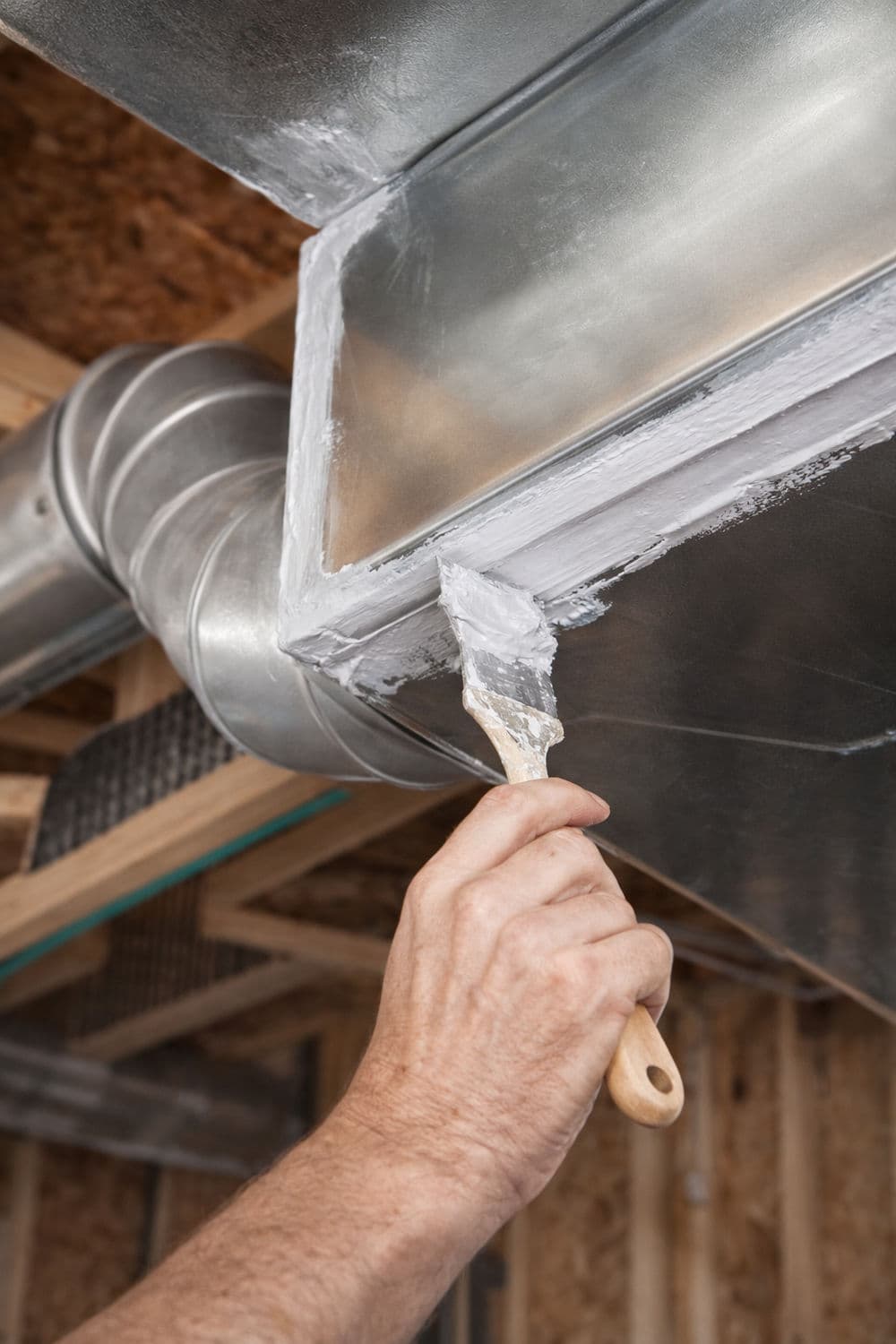 Person painting a metal duct in a basement renovation project with a putty knife.